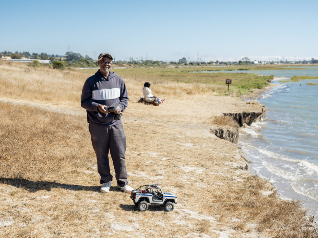 Ricky Thigpen stands on a beach with a black and white-striped hoodie and black pants, maneuvering a remote-control jeep.