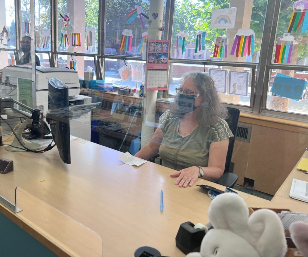 Alison Moreno sits in a black rolling chair at her desk with a computer, wearing a green shirt, glasses, and a black mask. She appears to be working at a Children's Library.