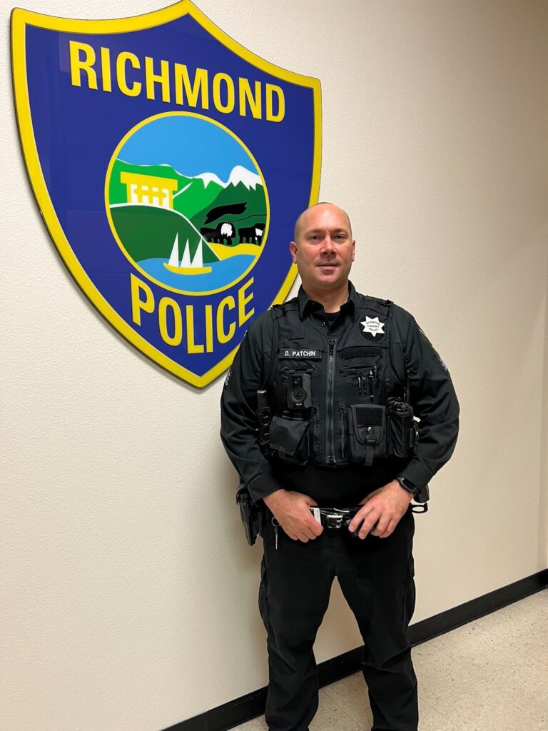 A police officer stands to the left of a blue and yellow Richmond Policie sign. He is bald, in blue uniform, his badge on his left chest, his nameplate D. Patchin on his right, his hands on his belt.