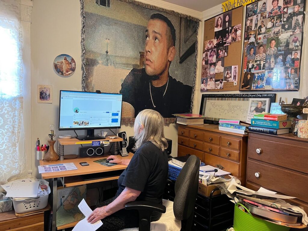 A gray-haired woman sits with her back to the camera, looking at a computer screen on a desk. Above the screen is a profile picture of a clean-shaven young man with short dark hair on a blanket. Hanging to her right iss a cork board fled with photo, mostly of a boy growing up.