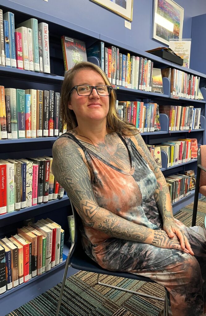 A woman in a multi-color matching top and pants and glasses, long blond hair, sits in the library in front of a stack of books.