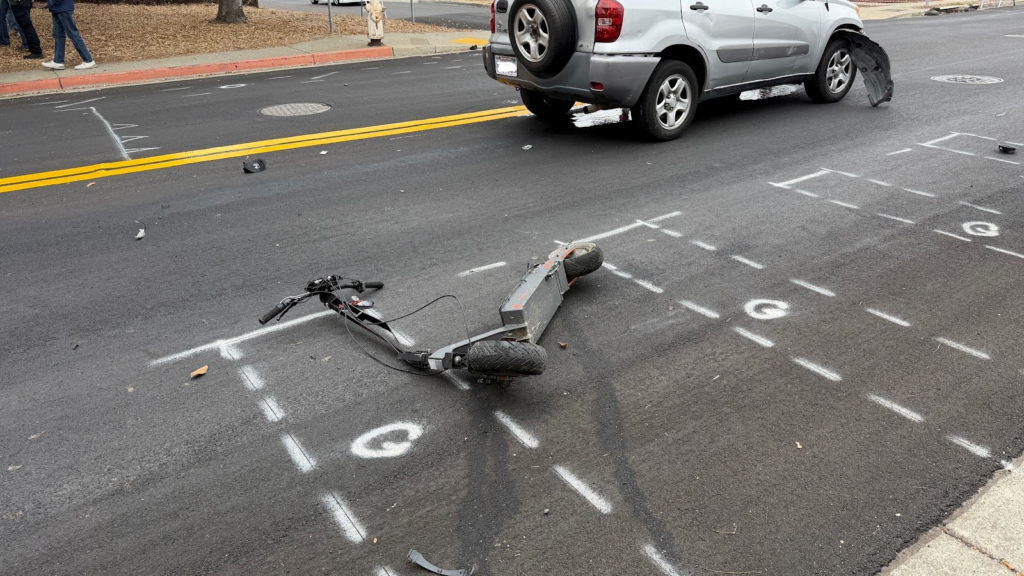 A mangled electric scooter sits on its side on a street with white chalk markings. Beside it is an SUV with its front tire shredded.
